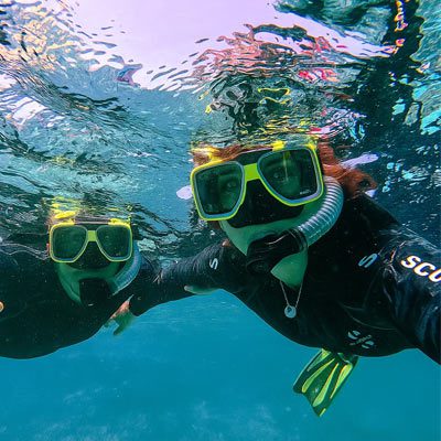 Two snorkelers underwater in tropical blue water, wearing neon masks and black wetsuits during a Miami reef tour.